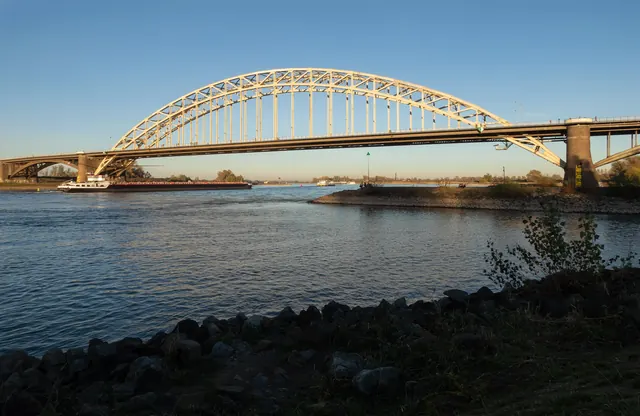 De Waalbrug over de Waal in de avondzon, met een vrachtschip eronder